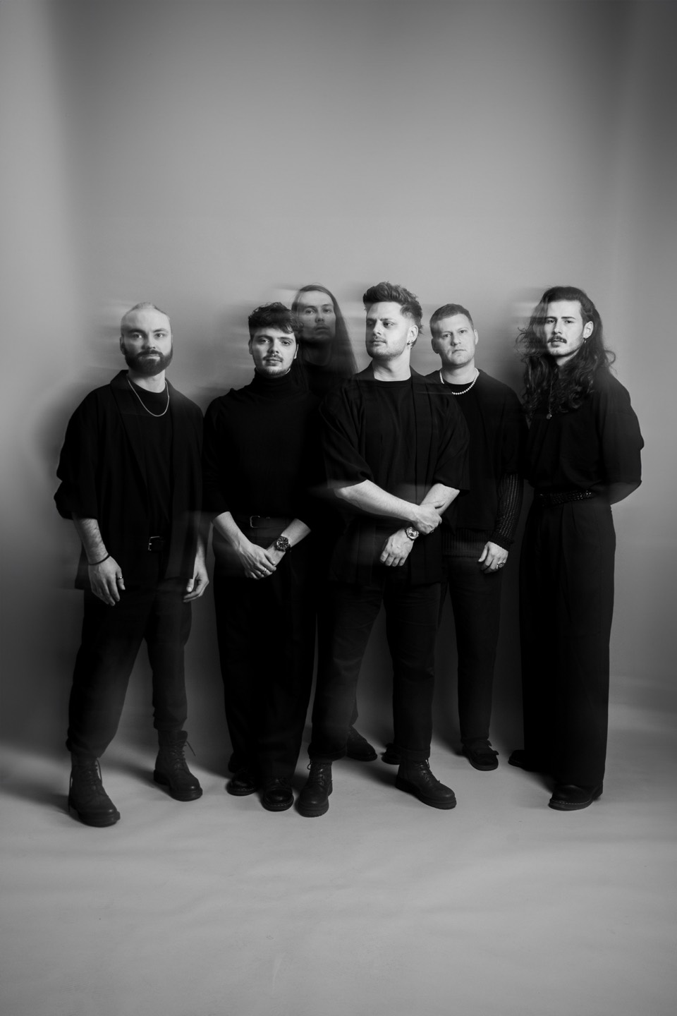 The six members of One Lunar Day standing against a pale wall, dressed in black, captured in a long exposure that blurs one figure into motion.