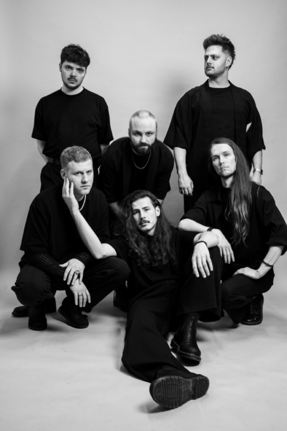 The six members of One Lunar Day arranged as a seated group portrait against a pale wall, all dressed in black.
